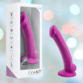 The Avant D9 Ergo MINI 6.5" Non-Porous Silicone Dildo in violet, featuring a curved shape, suction base, and dual density body-safe platinum silicone, stands beside its branded box. Soft pastel circles are blurred in the background.